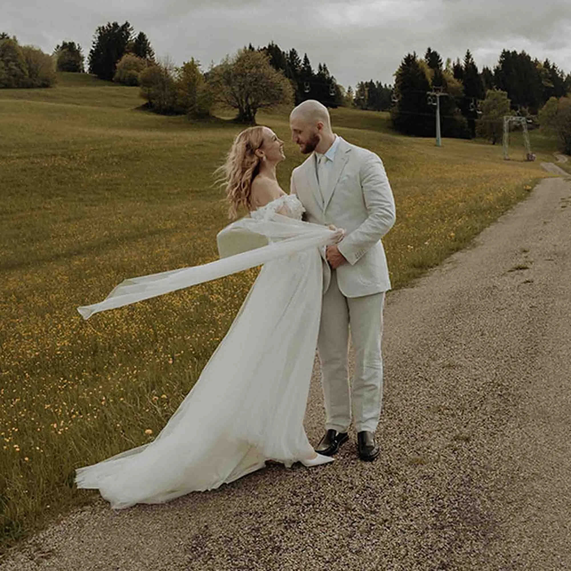 Braut und Bräutigam stehen auf einem Landweg inmitten einer grünen Wiese, während ihr Kleid und Schleier im Wind wehen.