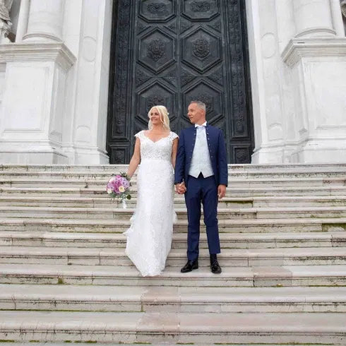Brautpaar steht Hand in Hand auf den Stufen einer Kirche, die Braut trägt ein elegantes Spitzenbrautkleid mit Blumenstrauß, der Bräutigam einen dunkelblauen Anzug vor einer großen Kirchentür.