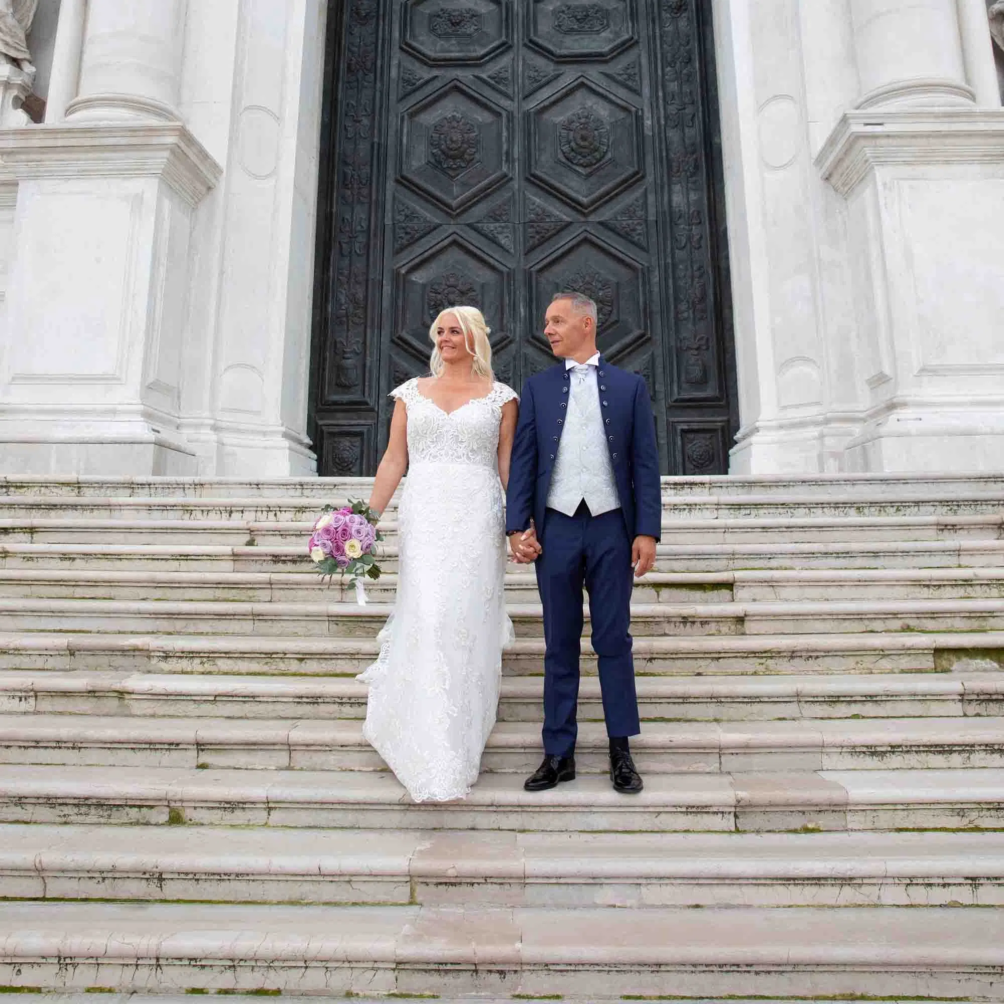 Brautpaar steht Hand in Hand auf den Stufen einer Kirche, die Braut trägt ein elegantes Spitzenbrautkleid mit Blumenstrauß, der Bräutigam einen dunkelblauen Anzug vor einer großen Kirchentür.