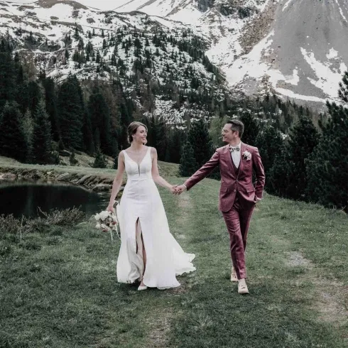Brautpaar hält sich an den Händen und spaziert in eleganten Hochzeitsoutfits durch eine alpine Berglandschaft mit See, Tannenwald und schneebedeckten Gipfeln im Hintergrund.