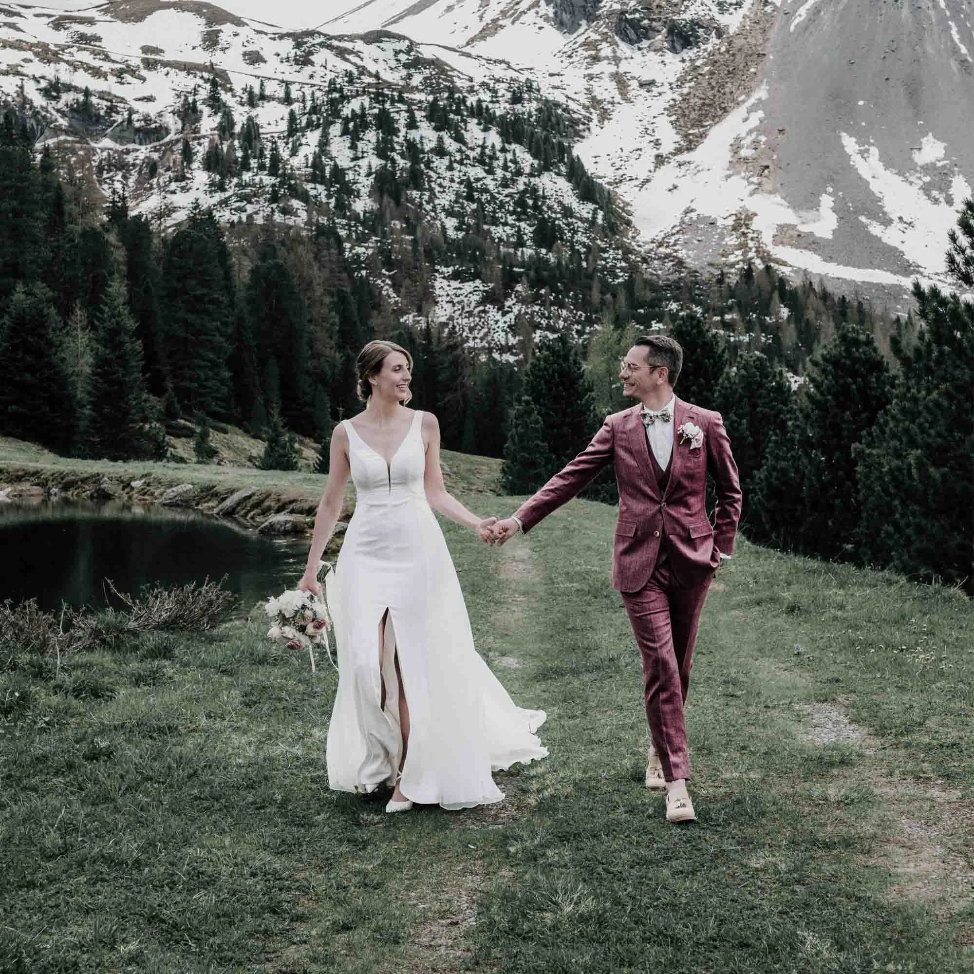 Brautpaar hält sich an den Händen und spaziert in eleganten Hochzeitsoutfits durch eine alpine Berglandschaft mit See, Tannenwald und schneebedeckten Gipfeln im Hintergrund.