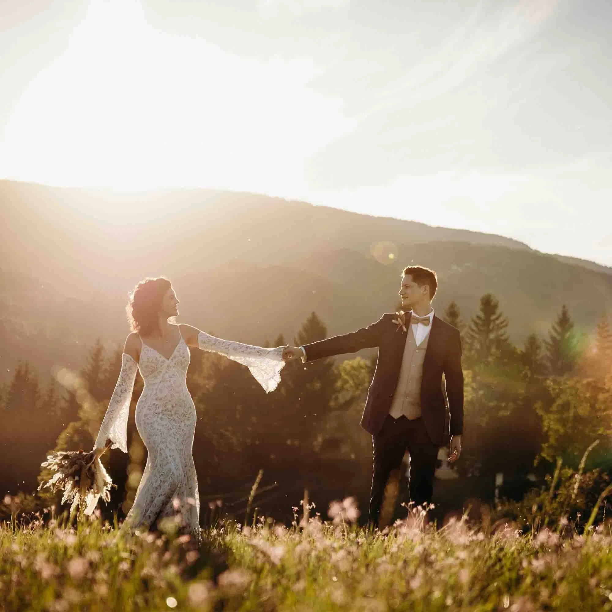 Brautpaar hält sich bei goldenem Abendlicht auf einer blühenden Wiese an den Händen, umgeben von Bergen und Natur – romantisches Hochzeitsshooting im Sonnenuntergang.