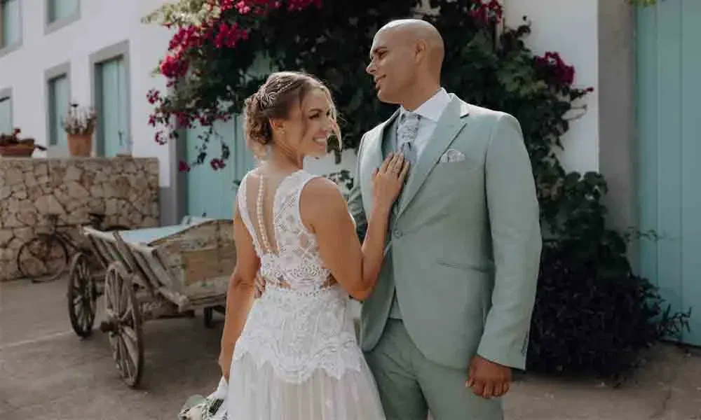 Braut und Bräutigam stehen lachend nebeneinander vor einer Steinmauer und blühenden Bougainvillea, während sie sich liebevoll ansehen.