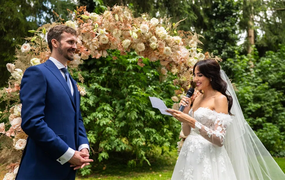 Emotionale freie Trauung im Grünen, bei der die Braut unter einem Blumenbogen ihr Eheversprechen vorliest und der Bräutigam aufmerksam zuhört.