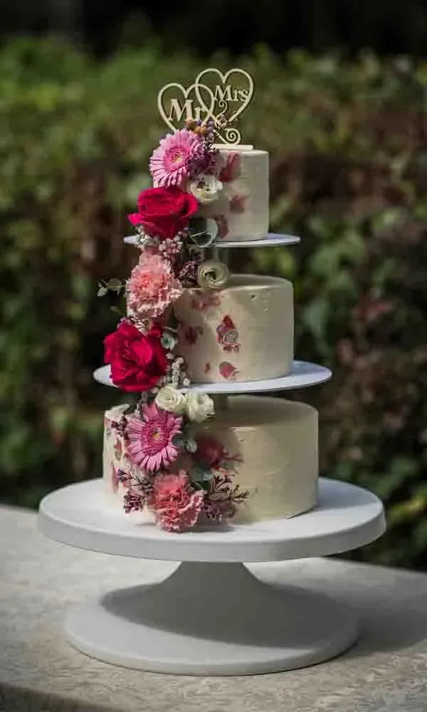 Dreistöckige Hochzeitstorte im Freien, dekoriert mit pinken und roten frischen Blumen sowie einem “Mr & Mrs”-Topper.