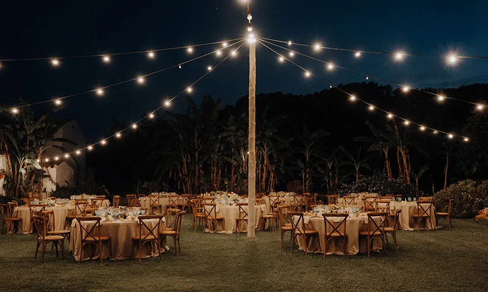 Stimmungsvoll beleuchtete Outdoor-Hochzeitstafel bei Nacht, mit runden Tischen unter einem Netz aus warmen Lichterketten.
