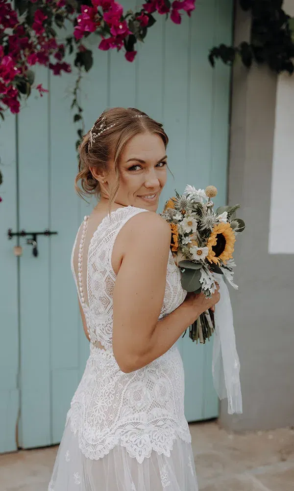 Lächelnde Braut im Spitzenkleid blickt über die Schulter und hält einen Strauß mit Sonnenblumen vor mintfarbener Tür.