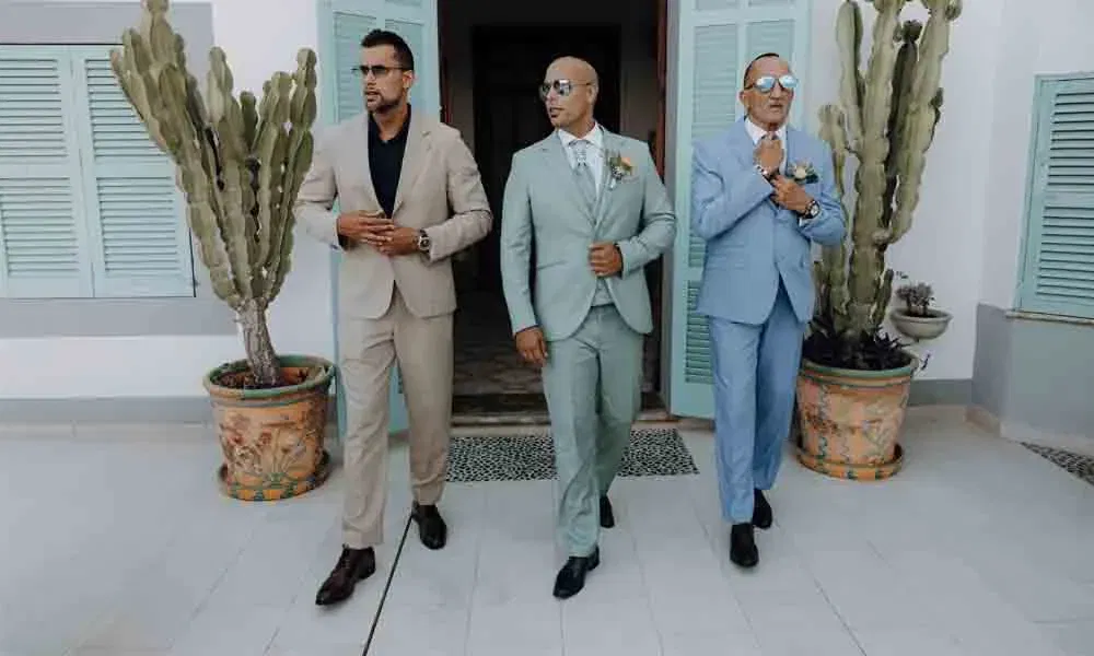 Bräutigam und zwei Männer in eleganten Anzügen treten selbstbewusst aus einer mintfarbenen Tür, flankiert von großen Kakteentöpfen.