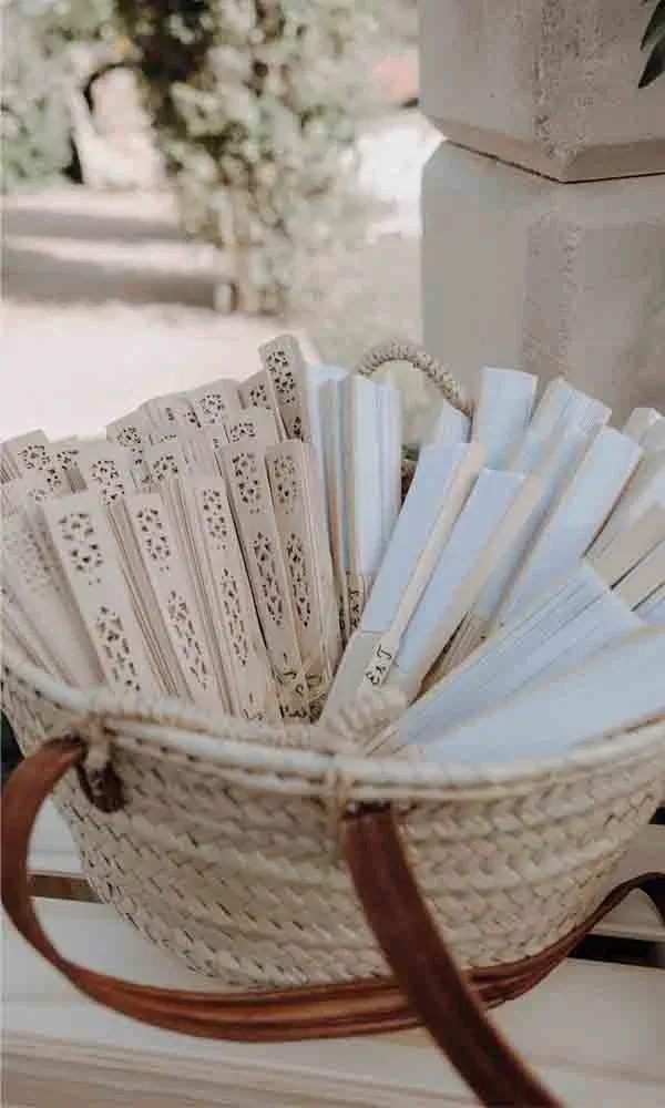 Geflochtene Korbtasche gefüllt mit dekorativen Holzfächern, bereitgestellt für eine sommerliche Hochzeit im Freien.
