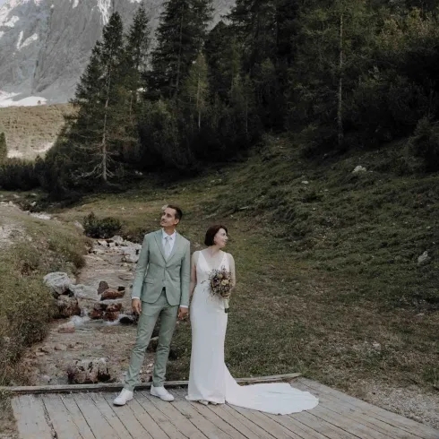 Braut und Bräutigam stehen Hand in Hand auf einem Holzsteg vor einer Berglandschaft, während ein Bach hinter ihnen entlangfließt.