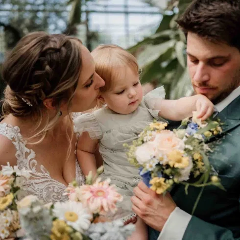 Inniger Hochzeitsmoment mit Kind: Braut und Bräutigam halten ihr kleines Kind im Arm, umgeben von floralen Sträußen und natürlicher Kulisse