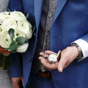 Detailaufnahme eines Bräutigams im blauen Anzug mit Taschenuhr an Kette, während die Braut einen weißen Blumenstrauß hält.