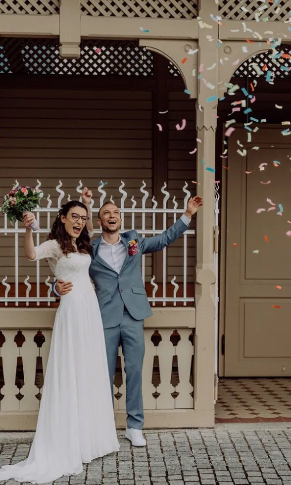 Frisch verheiratetes Brautpaar jubelt vor dem Standesamt, Konfetti fliegt durch die Luft, die Braut im schlichten Brautkleid mit Strauß, der Bräutigam im hellblauen Anzug.