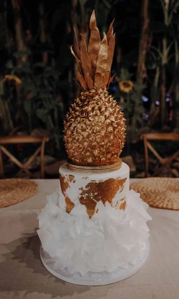 Hochzeitstorte mit weißem Rüschen-Design, goldener Weltkarten-Illustration und imposanter Ananas als goldfarbenem Topper.