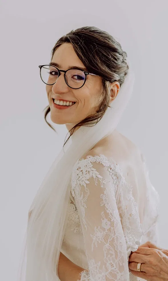 Braut mit hochgesteckter Frisur und Brille zeigt die Rückansicht ihres Brautkleides, feine Spitze am Rücken und langer Schleier, aufgenommen in einem sonnigen Garten.