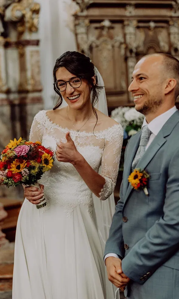 Lachende Braut zeigt den Daumen hoch in der Kirche, hält einen farbenfrohen Brautstrauß, der Bräutigam steht lächelnd neben ihr.