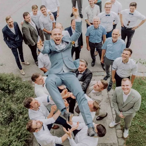 Bräutigam im hellblauen Anzug wird lachend von seinen Freunden in die Luft geworfen, umgeben von Hochzeitsgästen bei ausgelassener Stimmung im Freien.