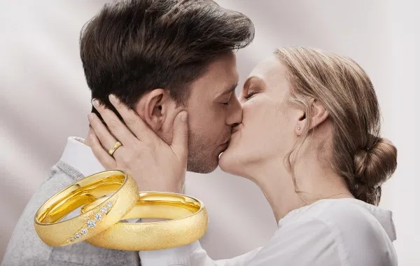 Ein verliebtes Brautpaar küsst sich innig, im Vordergrund sind zwei goldene Trauringe zu sehen, einer davon mit funkelnden Diamanten besetzt.
