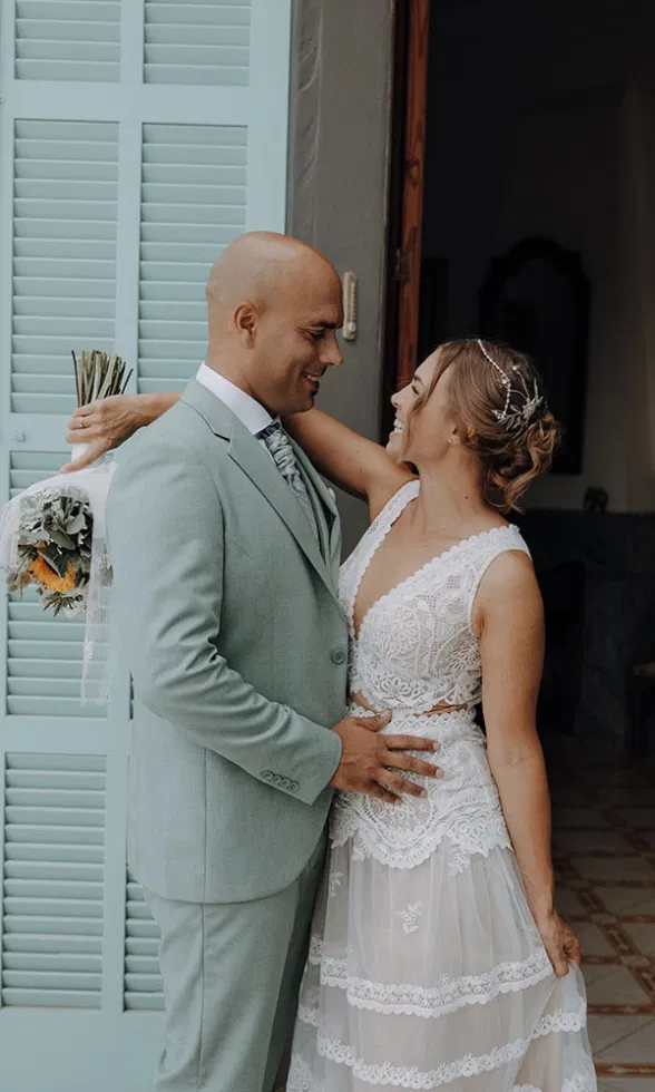 Braut und Bräutigam stehen sich liebevoll gegenüber in einer offenen Tür mit pastellgrünen Fensterläden, sie im detailreichen Spitzenbrautkleid mit Haarschmuck, er im hellgrünen Anzug, ein inniger Moment voller Nähe und Vorfreude.