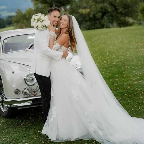 Brautpaar in eleganter Hochzeitsmode steht eng umschlungen vor einem weißen Oldtimer auf einer grünen Wiese, die Braut trägt ein zartes Tüllkleid mit floralen Details und langen Schleier.
