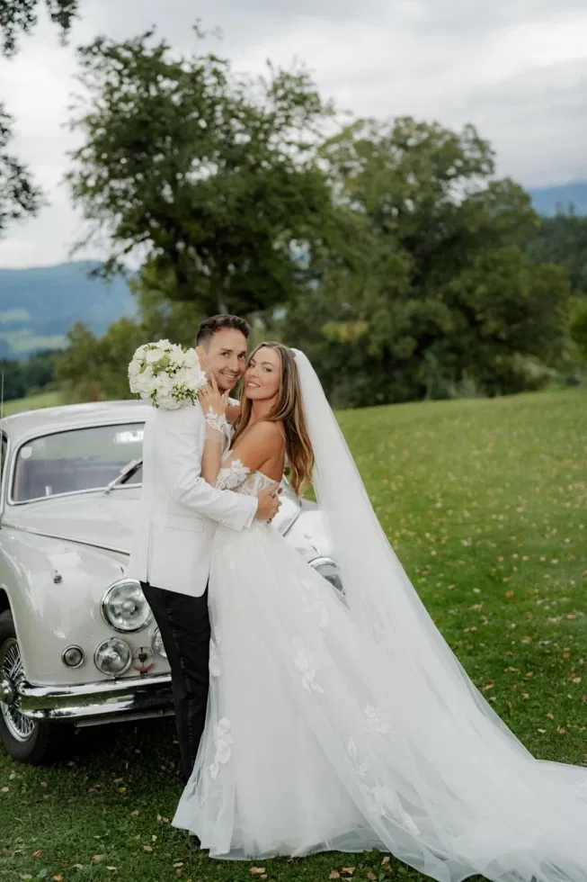 Brautpaar umarmt sich liebevoll vor einem weißen Oldtimer auf einer grünen Wiese, die Braut in einem floralen Brautkleid mit langem Schleier, der Bräutigam im eleganten Smoking, umgeben von Natur und sanfter Berglandschaft.