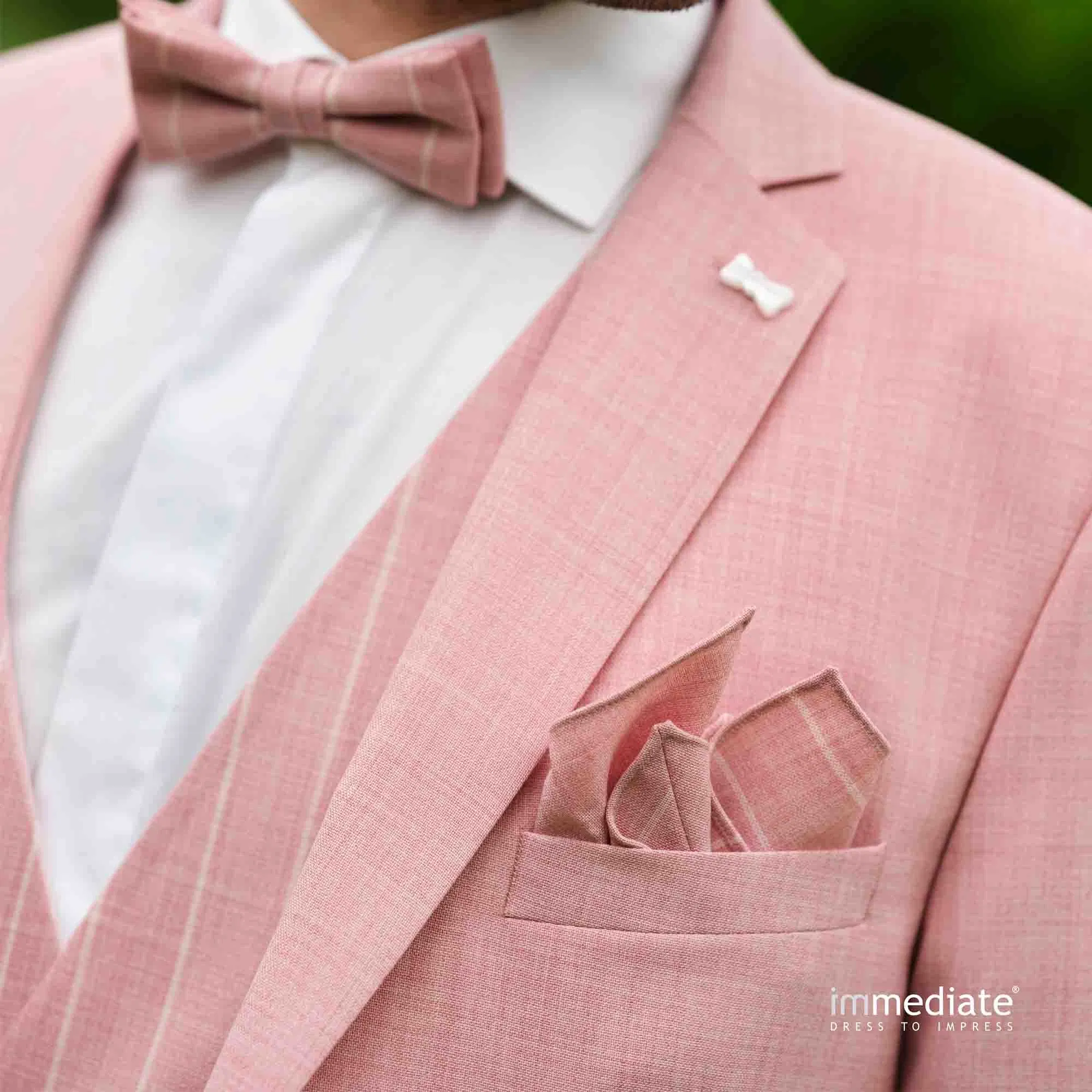 Detailaufnahme eines Bräutigams im roséfarbenen Anzug mit Fliege, Einstecktuch und feiner Reversnadel.