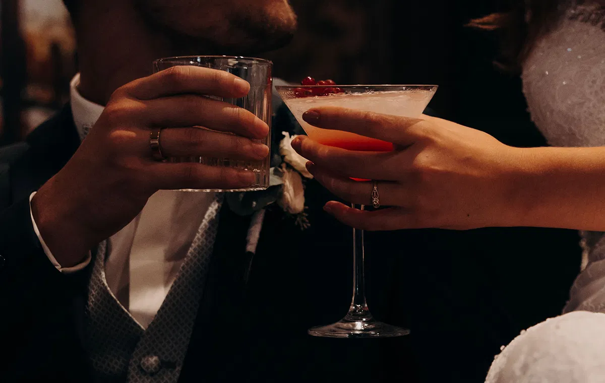 Braut und Bräutigam stoßen mit einem roten Cocktail und einem Whiskyglas bei stimmungsvoller Beleuchtung an.