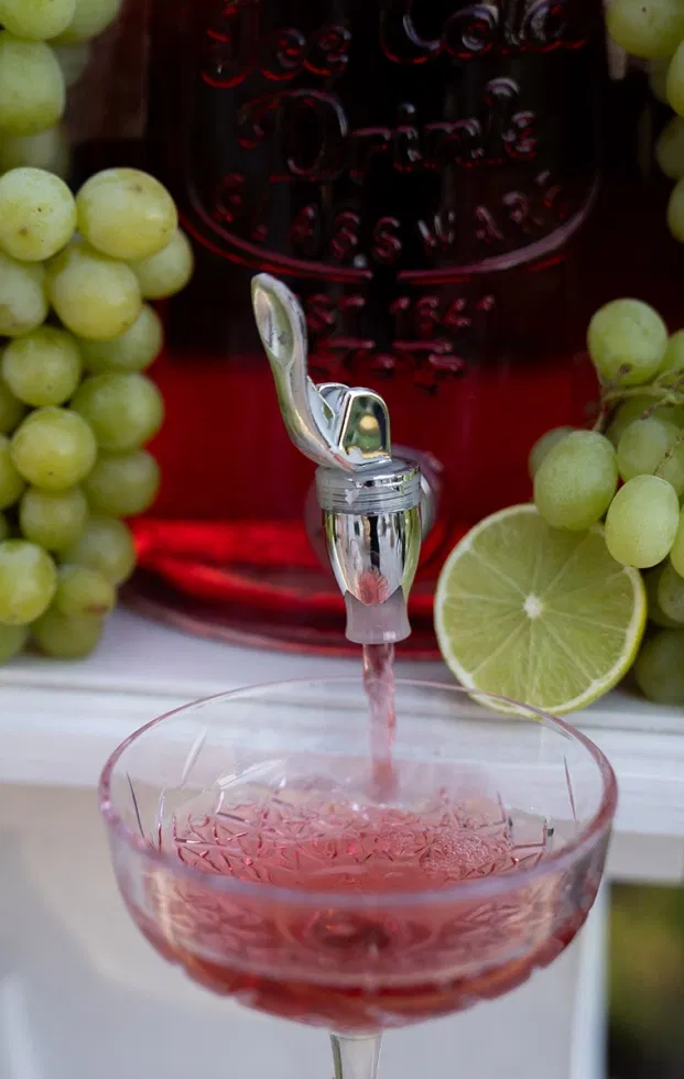 Roséfarbener Signature-Drink fließt aus einem Getränkespender in ein Glas, dekoriert mit grünen Trauben und Limette.