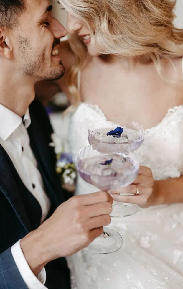 Lächelndes Hochzeitspaar stößt mit zwei lilafarbenen Signature-Cocktails inklusive essbarer Blüte an
