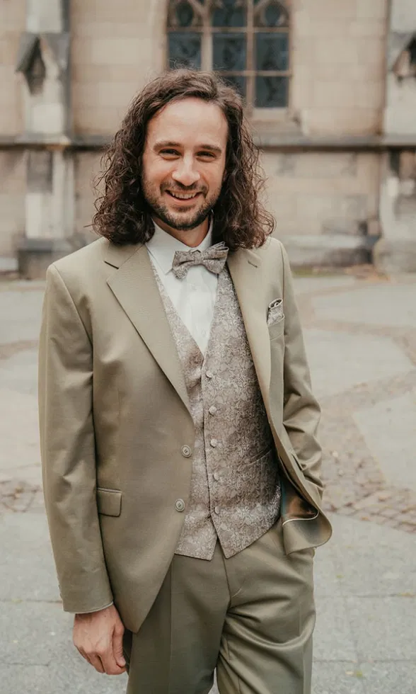 Bräutigam mit lockigem Haar im beigen Anzug mit gemusterter Weste und Fliege, entspanntes Porträt vor historischer Steinarchitektur.