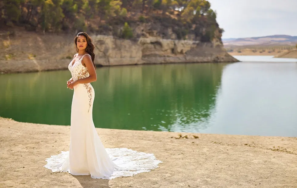 Figurbetontes Brautkleid mit Spitzendetails und fließender Schleppe, inszeniert an einem ruhigen See in weiter Naturlandschaft.