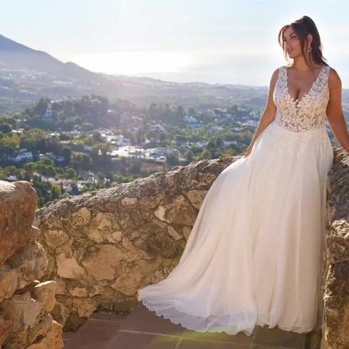 Braut im fließenden A-Linien-Brautkleid mit Spitzenoberteil, inszeniert auf einer Natursteinmauer mit mediterraner Landschaft im Hintergrund.