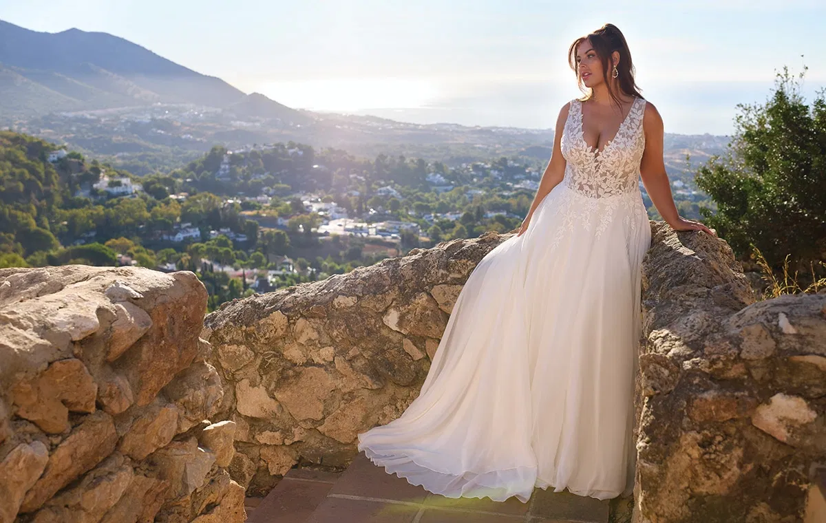 Braut im fließenden A-Linien-Brautkleid mit Spitzenoberteil, inszeniert auf einer Natursteinmauer mit mediterraner Landschaft im Hintergrund.