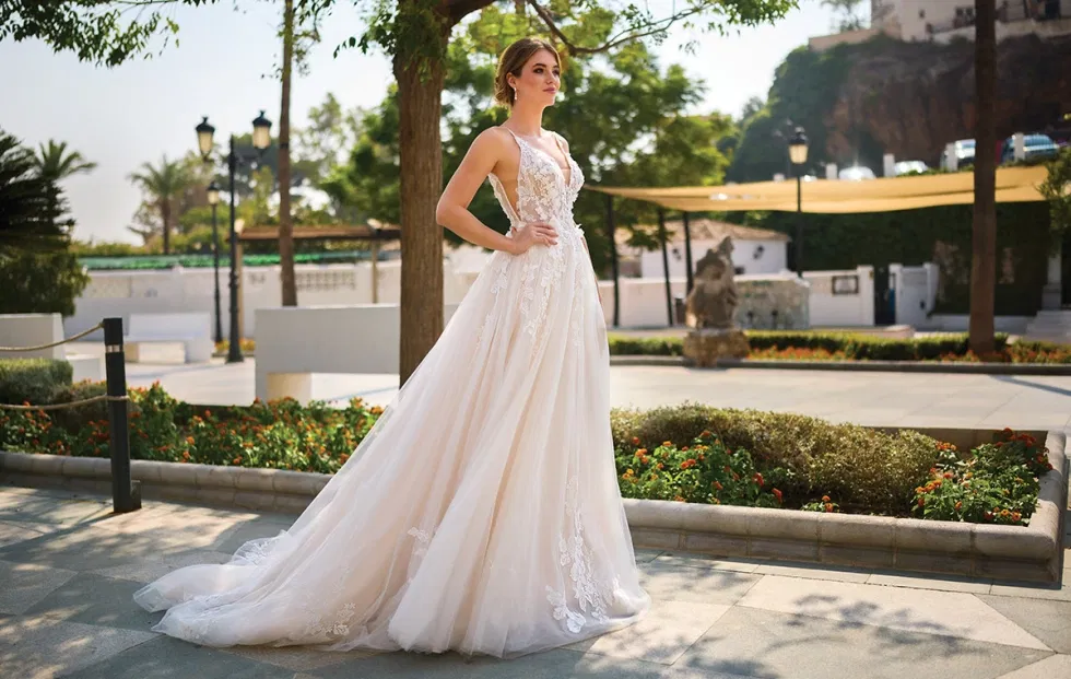 Elegantes Brautkleid mit tiefem V-Ausschnitt und transparenter Spitze, aufgenommen in einer gepflegten Gartenanlage mit klassischer Architektur.