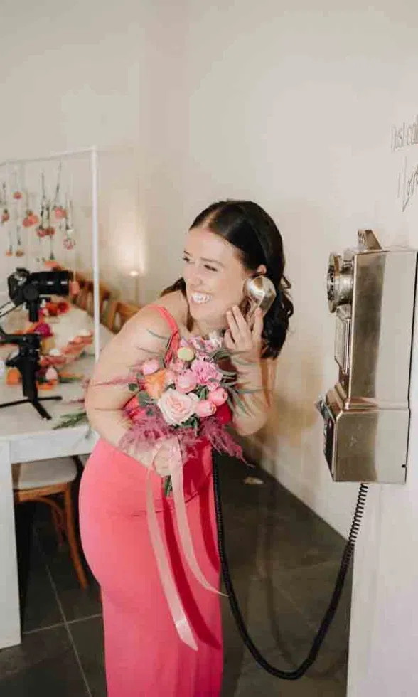 Brautjungfer im korallfarbenen Kleid hält einen Blumenstrauß und lächelt, während sie spielerisch an einem nostalgischen Wandtelefon lauscht, in einem hellen, modern dekorierten Hochzeitsambiente.