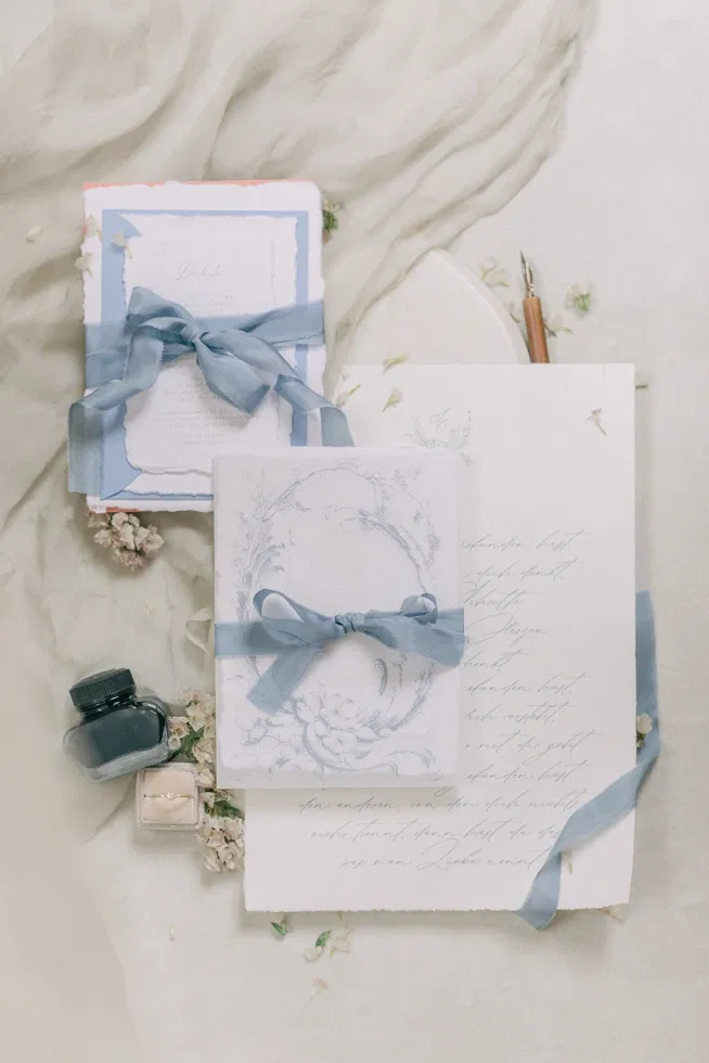 Edle Hochzeits­papeterie im Fine-Art-Stil mit handgeschriebenem Brief, Umschlägen, Seidenband in Staubblau, Tintenfass und Ringdetail auf hellem Stoff arrangiert.