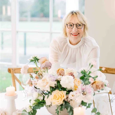 Wedding-Planerin dekoriert einen Hochzeitstisch mit üppigem Blumenarrangement in zarten Pastelltönen und Kerzen in hellem, lichtdurchflutetem Raum.