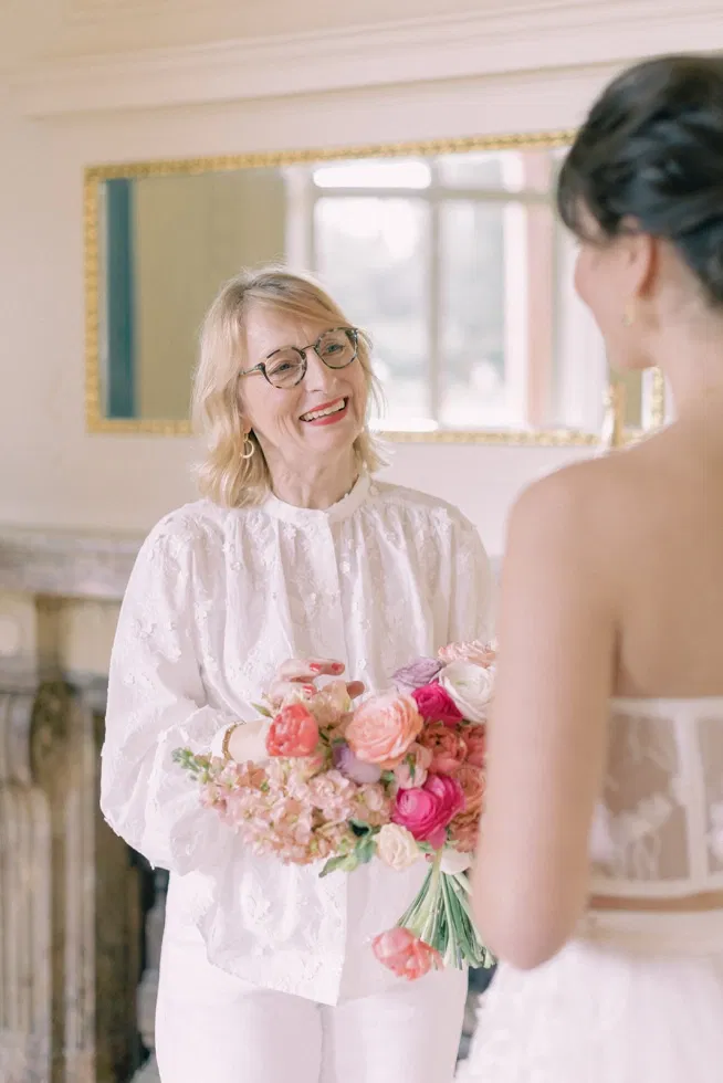 Wedding-Planerin überreicht einer Braut im hellen Innenraum einen üppigen Brautstrauß in Rosé- und Pastelltönen und lächelt sie herzlich an.