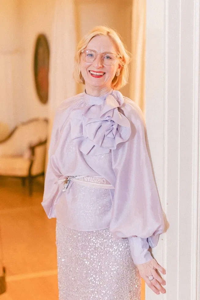 Porträt einer lächelnden Wedding-Planerin in fließendem, lavendelfarbenem Kleid mit feinen Details, aufgenommen in warmem Innenraumlicht.