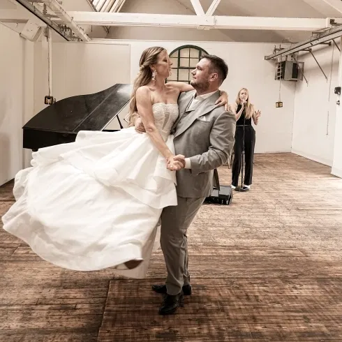 Brautpaar beim Hochzeitstanz in einer lichtdurchfluteten Location mit Holzboden: Der Bräutigam wirbelt die Braut im weißen, schwingenden Brautkleid über die Tanzfläche, während im Hintergrund eine Sängerin live begleitet – emotionaler Moment voller Nähe und Bewegung.
