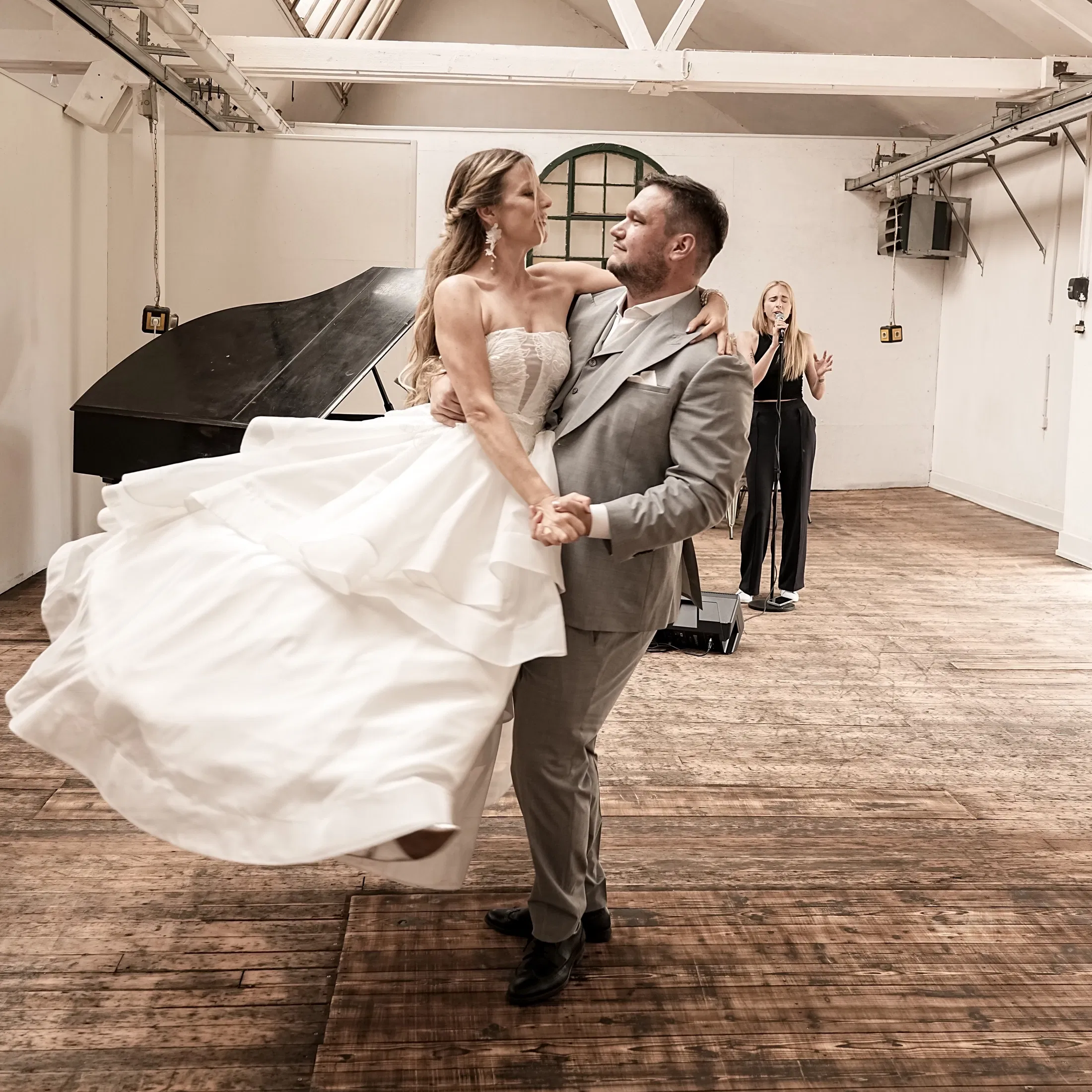 Brautpaar beim Hochzeitstanz in einer lichtdurchfluteten Location mit Holzboden: Der Bräutigam wirbelt die Braut im weißen, schwingenden Brautkleid über die Tanzfläche, während im Hintergrund eine Sängerin live begleitet – emotionaler Moment voller Nähe und Bewegung.
