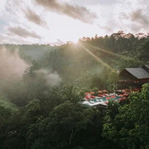 Luxuriöses Resort mit Infinity-Pool und roten Sonnenschirmen, eingebettet in eine üppige tropische Landschaft bei Sonnenaufgang mit Nebelschwaden über dem Dschungel.