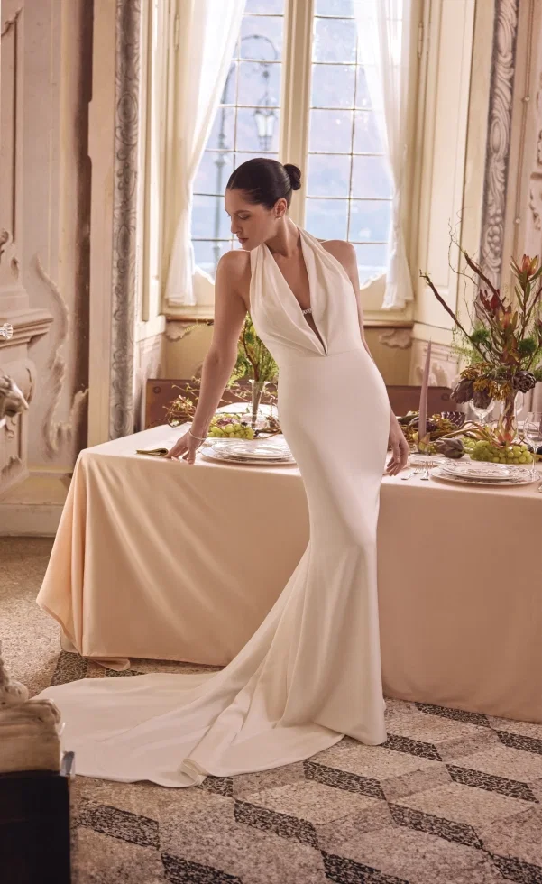 Braut in einem eleganten, figurbetonten Kleid mit tiefem Ausschnitt lehnt an einem festlich gedeckten Tisch in einem lichtdurchfluteten, historischen Raum.