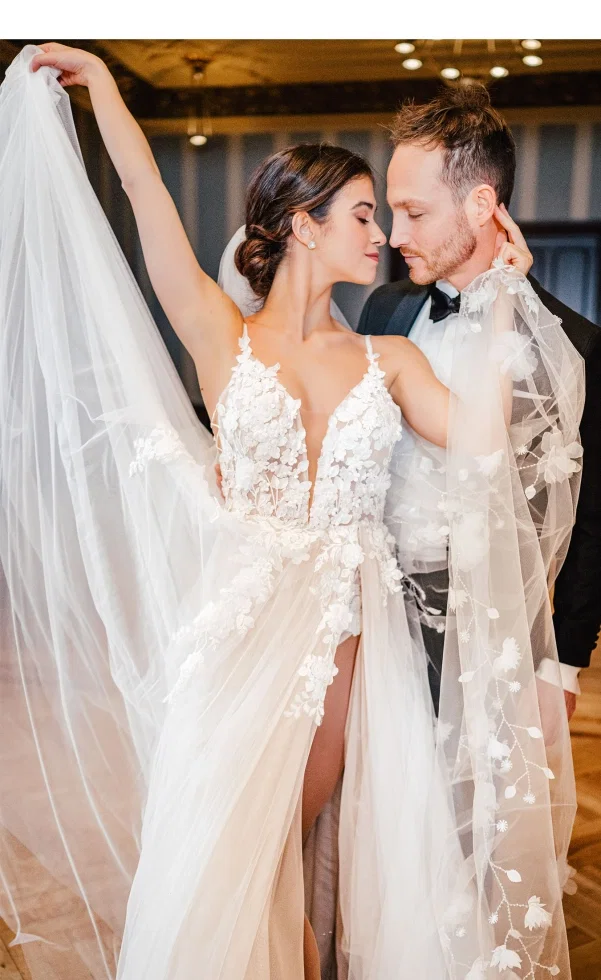 Elegantes Brautpaar in inniger Pose, die Braut hebt ihren Schleier an und trägt ein detailreiches Spitzenkleid mit tiefem Ausschnitt, während der Bräutigam sie nah bei sich hält.