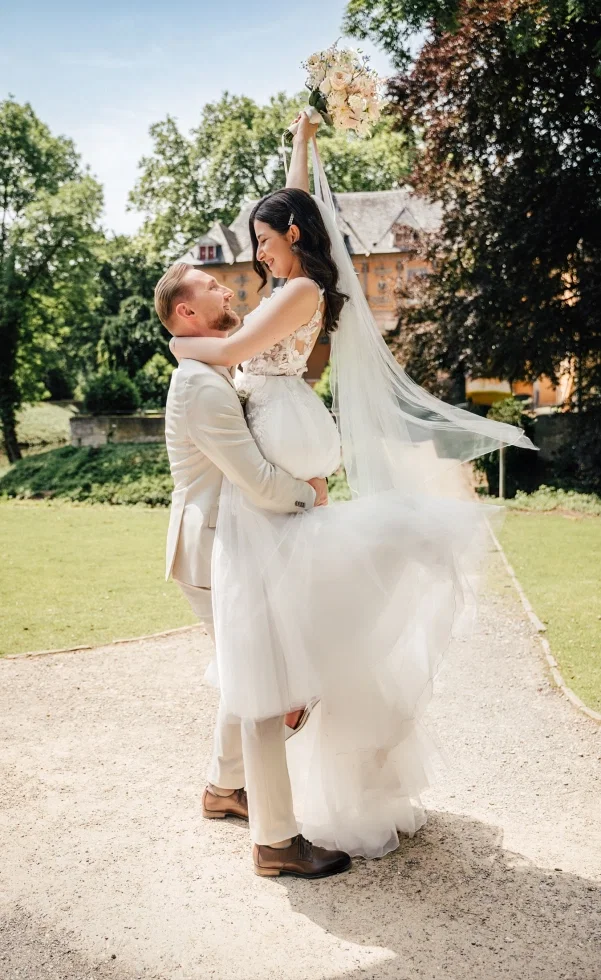 Glückliches Brautpaar im Park: Der Bräutigam hebt die Braut hoch, während sie ihren Blumenstrauß in die Luft streckt und sich strahlend ansehen