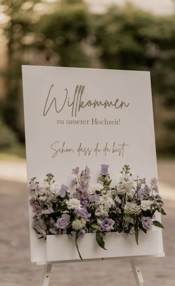 Elegantes Hochzeits-Willkommensschild auf einer Staffelei mit floraler Dekoration in Weiß und Flieder, liebevolle Begrüßung der Gäste im Außenbereich