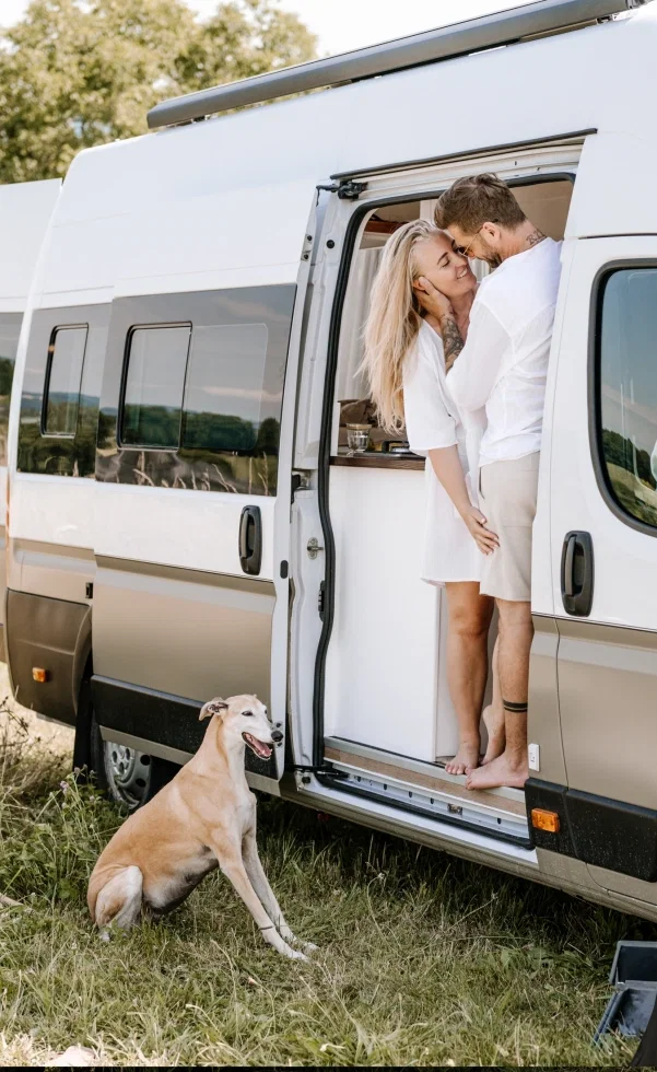 Verliebtes Paar steht eng umschlungen in der Tür eines Vans, während ihr Hund davor im Gras sitzt und aufmerksam zuschaut