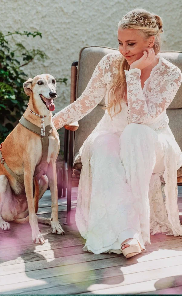 Braut im Spitzenkleid sitzt entspannt auf einer Terrasse und streichelt liebevoll ihren Hund, der mit Halsband und Geschirr neben ihr sitzt