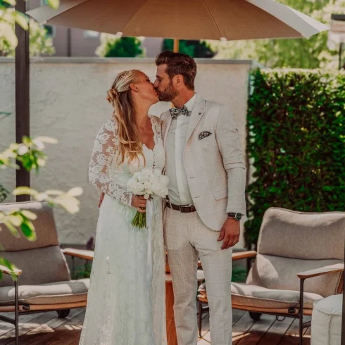 Brautpaar küsst sich unter einem Sonnenschirm im Garten – sie in langärmligem Spitzenkleid mit Brautstrauß, er in hellem Anzug mit Fliege, umgeben von sommerlicher Lounge-Atmosphäre