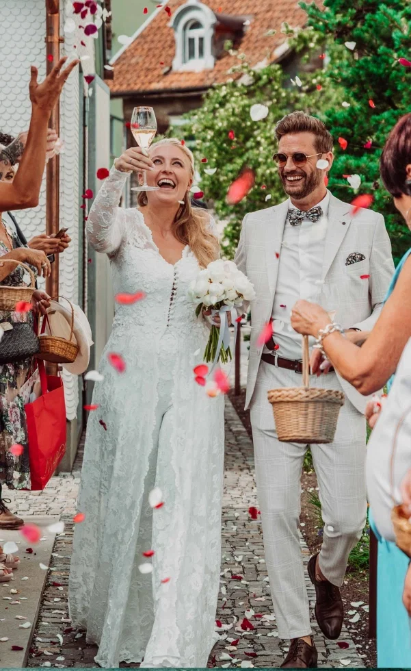 Fröhliches Brautpaar läuft nach der Trauung durch eine Gasse, während Gäste rote und weiße Blütenblätter werfen – die Braut hebt lachend ein Glas, der Bräutigam strahlt neben ihr